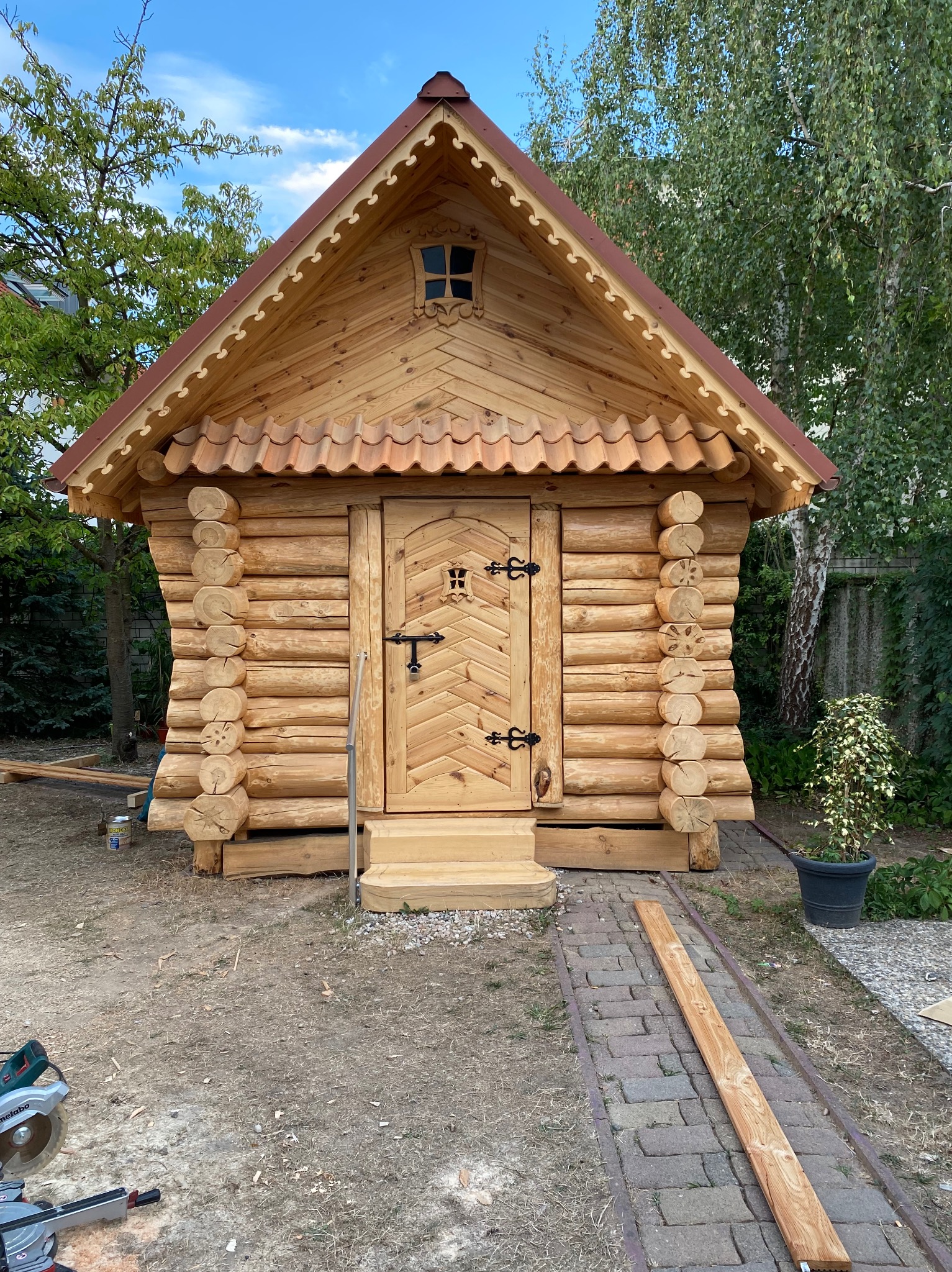 Mała, drewniana konstrukcja z bali, przypominająca domek, z dekoracyjnymi wykończeniami dachu i drzwi, umieszczona w ogrodzie, widoczna piła tarczowa i deski sugerują etap budowy.