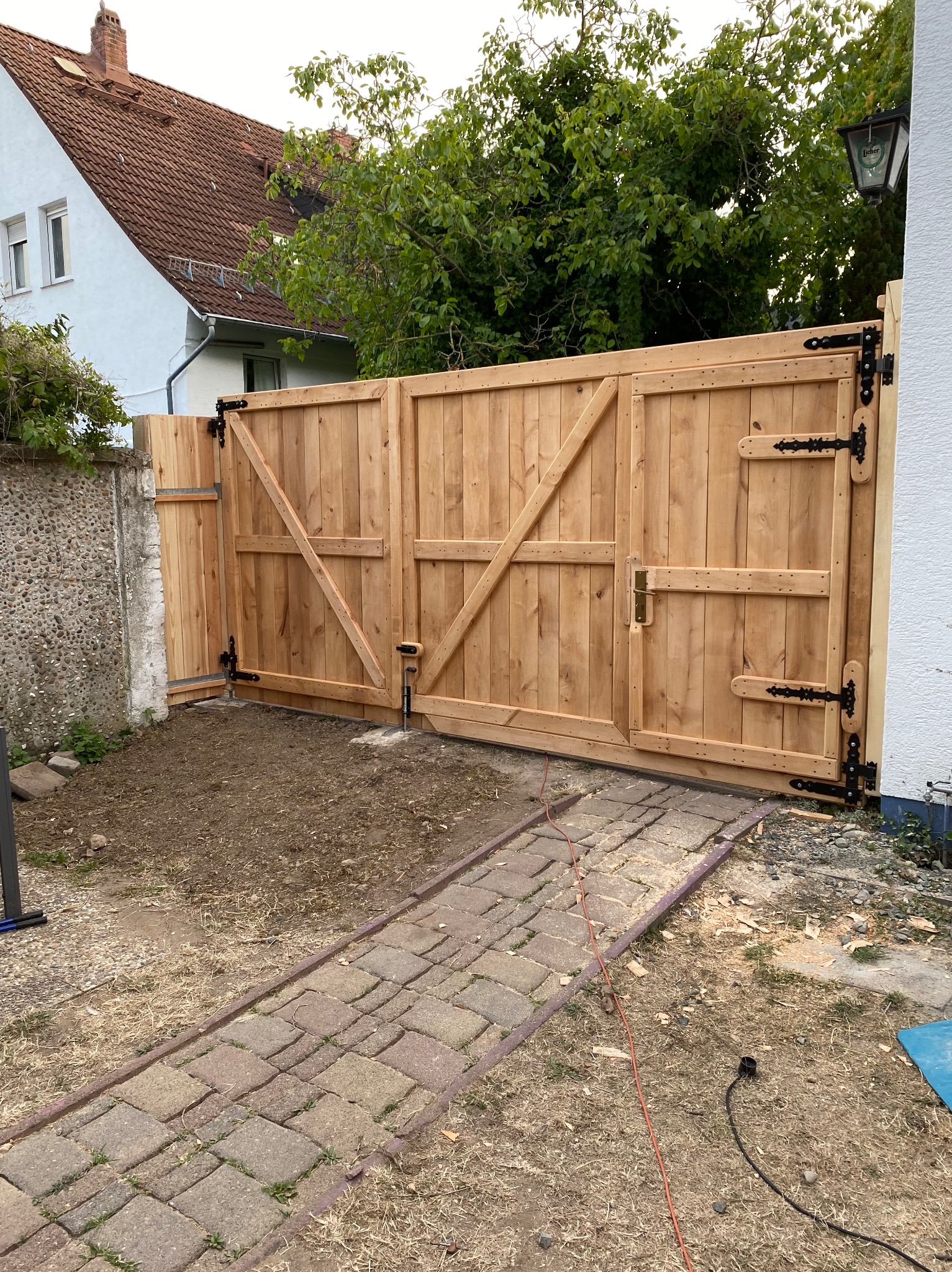 Nowa, dwuskrzydłowa brama z jasnego drewna z czarnymi okuciami, zamontowana przy kamiennym murze i białym budynku, widoczna ścieżka z kostki brukowej i resztki po montażu.