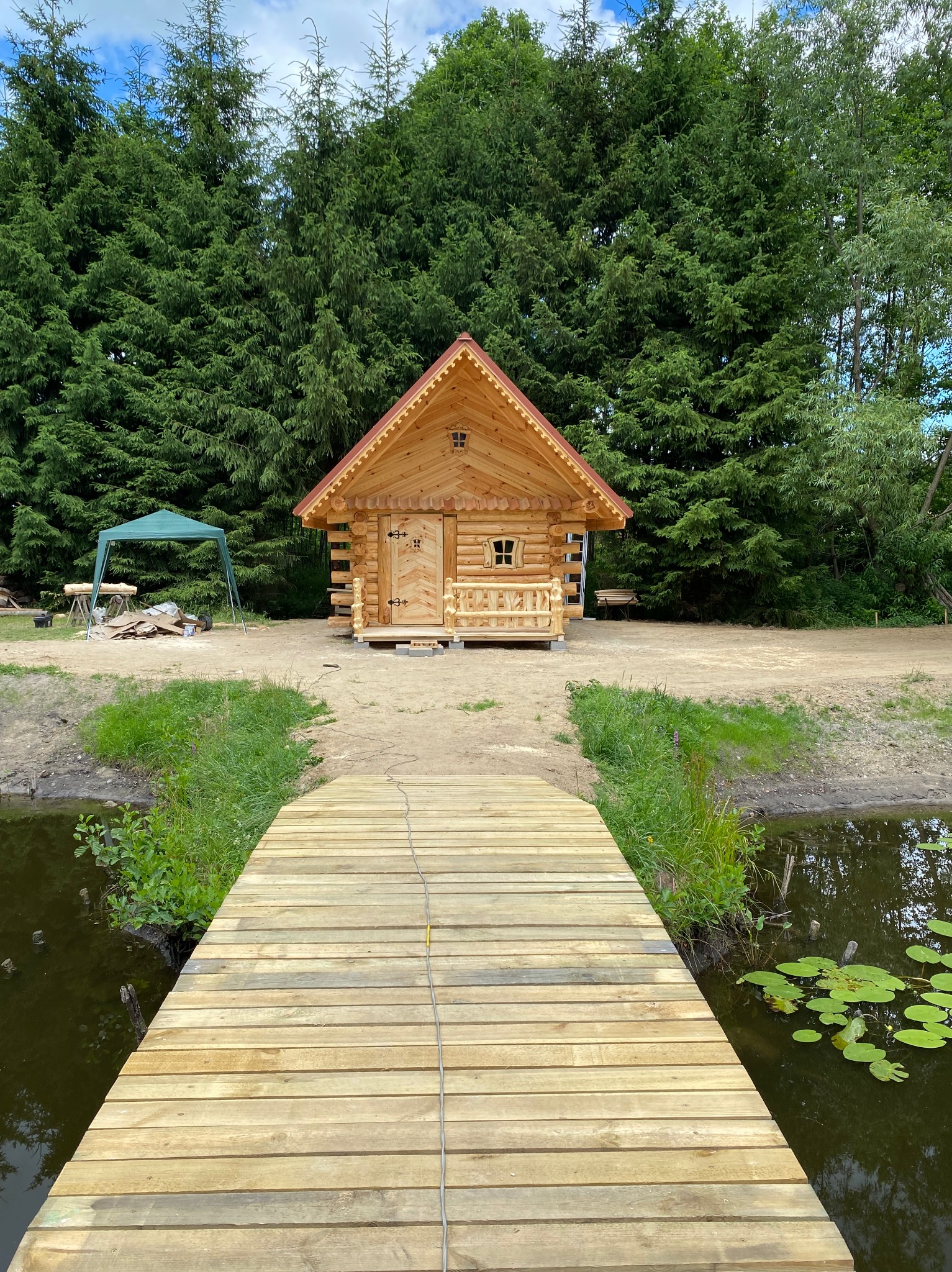 Nowo wybudowana, drewniana sauna z bali, z małą werandą, widoczna z drewnianego pomostu nad stawem, w tle gęsty las iglasty i rozłożony namiot.