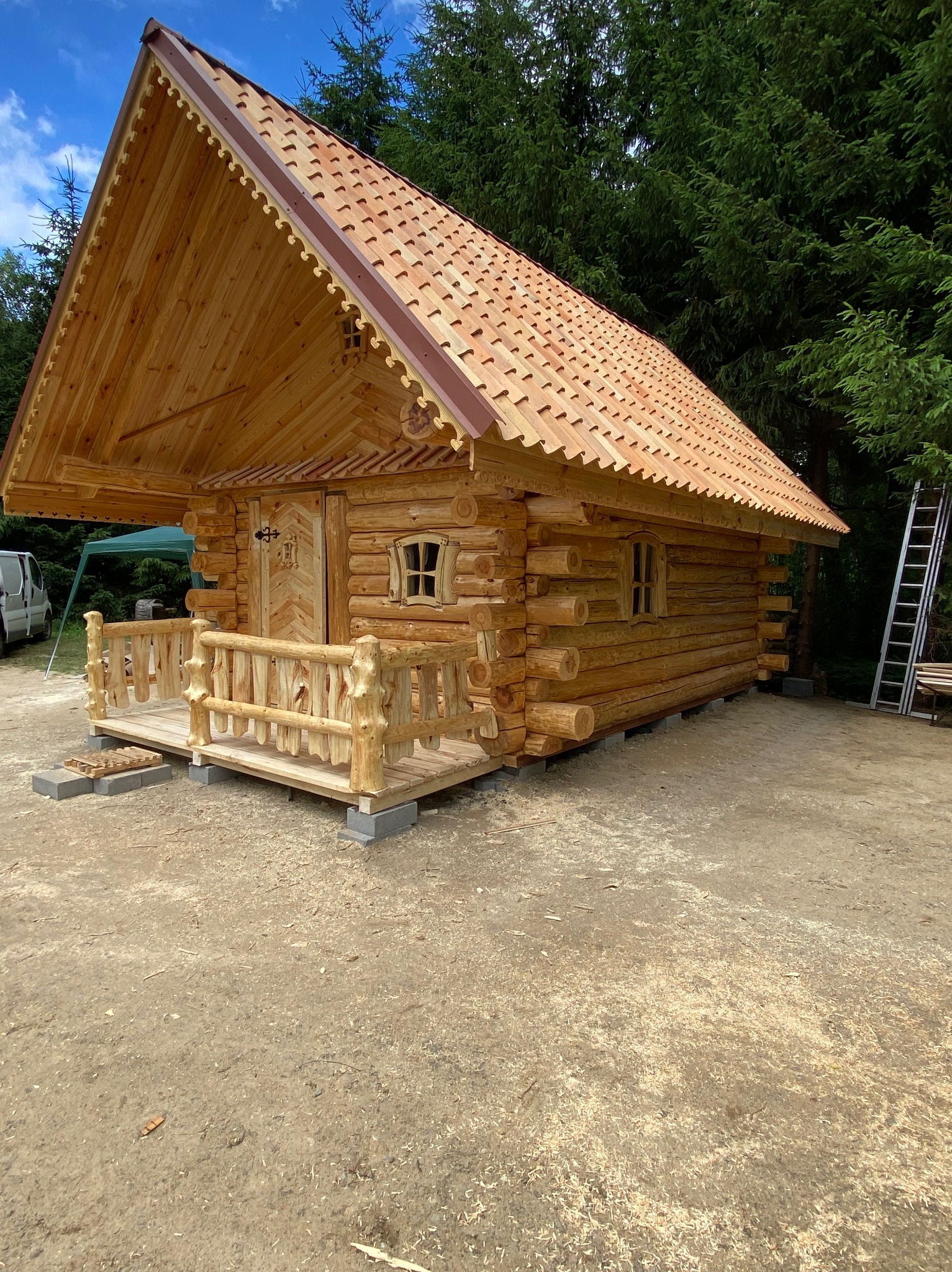 Drewniana konstrukcja z bali, prawdopodobnie domek letniskowy lub mała sauna, z ozdobnym dachem i małym tarasem z balustradą, umieszczona na betonowych fundamentach. W tle widoczne drzewa i drabina.