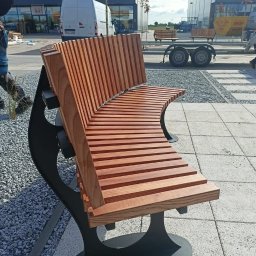 Krystian Kucz - Nowoczesna ławka parkowa z drewnianym siedziskiem i oparciem ułożonym z listew, na czarnej metalowej konstrukcji, umieszczona na placu wyłożonym kostką i żwirem, w tle budynek sklepu.