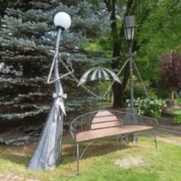 Oryginalna metalowa rzeźba parkowa przedstawiająca dwie stylizowane latarnie i parasol, tworzące kompozycję nad ławką z drewnianym siedziskiem, na tle zieleni parku.