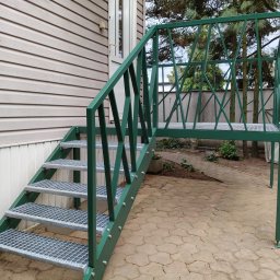 Krystian Kucz - Zielone schody metalowe z ażurową balustradą prowadzą do drzwi domu z sidingiem. Widoczne mocowania śrubowe i ażurowa konstrukcja balustrady. Tło stanowią drzewa i kostka brukowa.