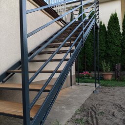 Krystian Kucz - Zewnętrzne schody metalowe z drewnianymi stopniami, prowadzące na balkon. Konstrukcja w kolorze grafitowym, tło stanowią tuje i elewacja budynku.