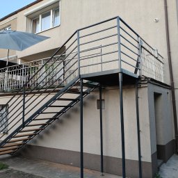Krystian Kucz - Zewnętrzne schody metalowe z drewnianymi stopniami, prowadzące na balkon w Mieścisko. Konstrukcja pomalowana na czarno, widok z dołu, jasne tło budynku.