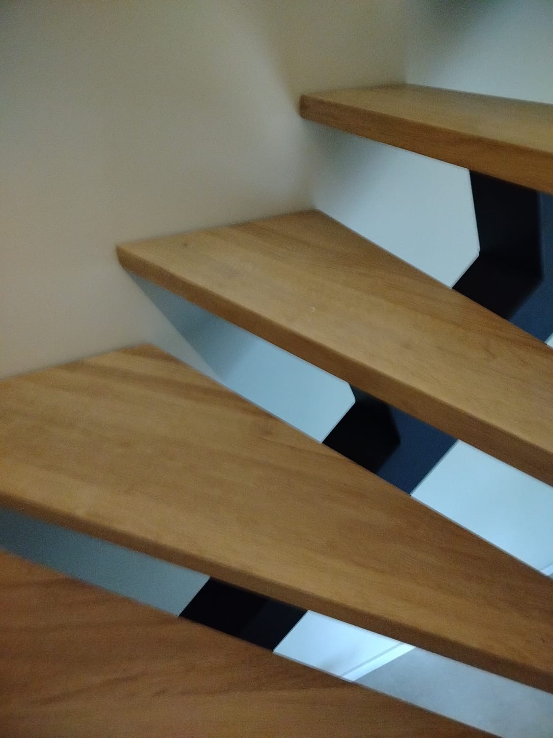 Fragment schodów z drewnianymi stopniami o ciepłym odcieniu, wspartych na konstrukcji z białych i czarnych elementów, przylegających do jasnej ściany.