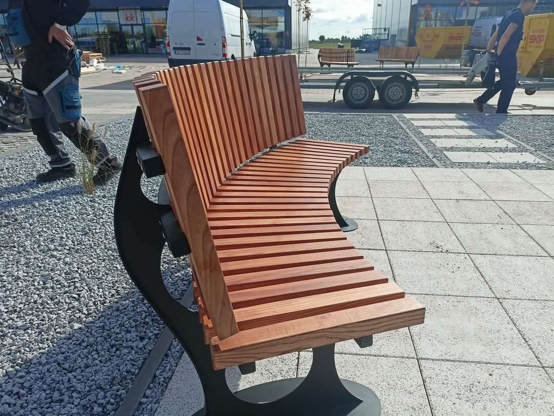 Nowoczesna ławka parkowa z drewnianym siedziskiem i oparciem ułożonym z listew, na czarnej metalowej konstrukcji, umieszczona na placu wyłożonym kostką i żwirem, w tle budynek sklepu.