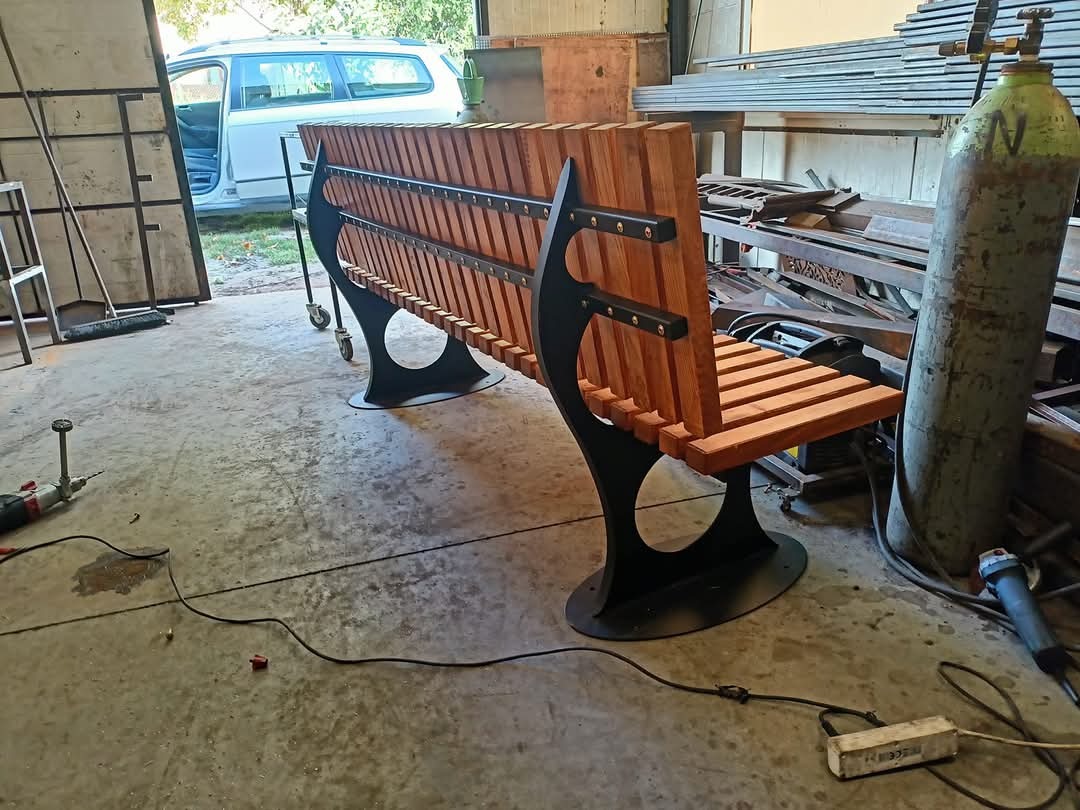 Nowoczesna ławka parkowa z drewnianym siedziskiem i czarną, metalową konstrukcją, widoczna w warsztacie z narzędziami i butlą spawalniczą w tle.