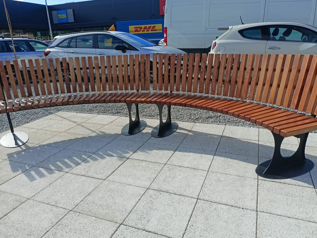 Zakrzywiona ławka z drewnianym siedziskiem i oparciem z pionowych listew, osadzona na czarnych, metalowych nogach o owalnej podstawie, na tle parkingu z samochodami i budynkami.