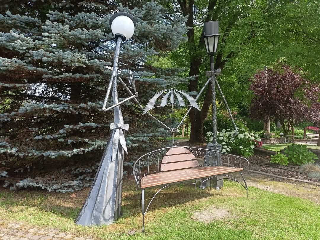 Oryginalna metalowa rzeźba parkowa przedstawiająca dwie stylizowane latarnie i parasol, tworzące kompozycję nad ławką z drewnianym siedziskiem, na tle zieleni parku.