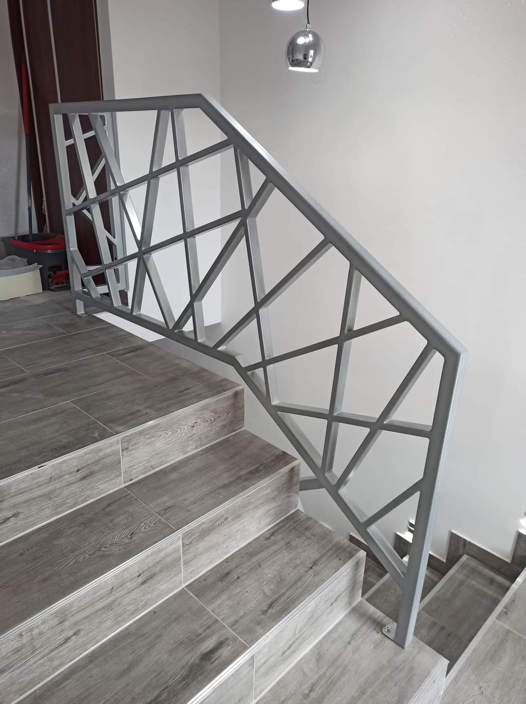 Nowoczesna, geometryczna balustrada metalowa w kolorze szarym, zamontowana na schodach wyłożonych płytkami imitującymi drewno, oświetlona przez wiszącą lampę w kształcie kuli.