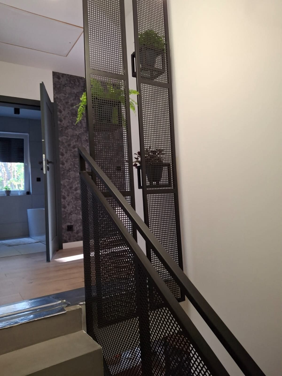 Nowoczesne schody metalowe z ażurową balustradą i wbudowanymi kwietnikami, widok z dołu, jasne wnętrze z elementami szarości i drewna.