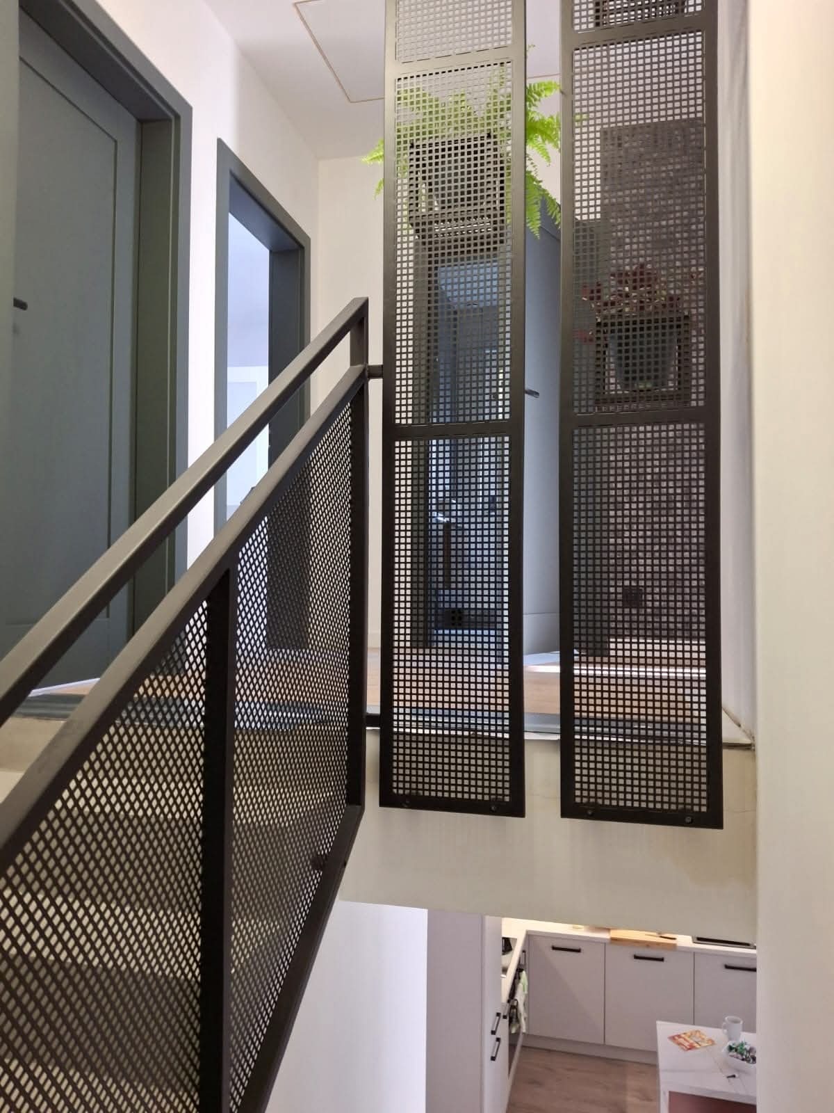Nowoczesna balustrada schodowa z metalowej siatki w ciemnym kolorze, z roślinami w tle. Minimalistyczny design, widok na dół schodów i fragment kuchni.