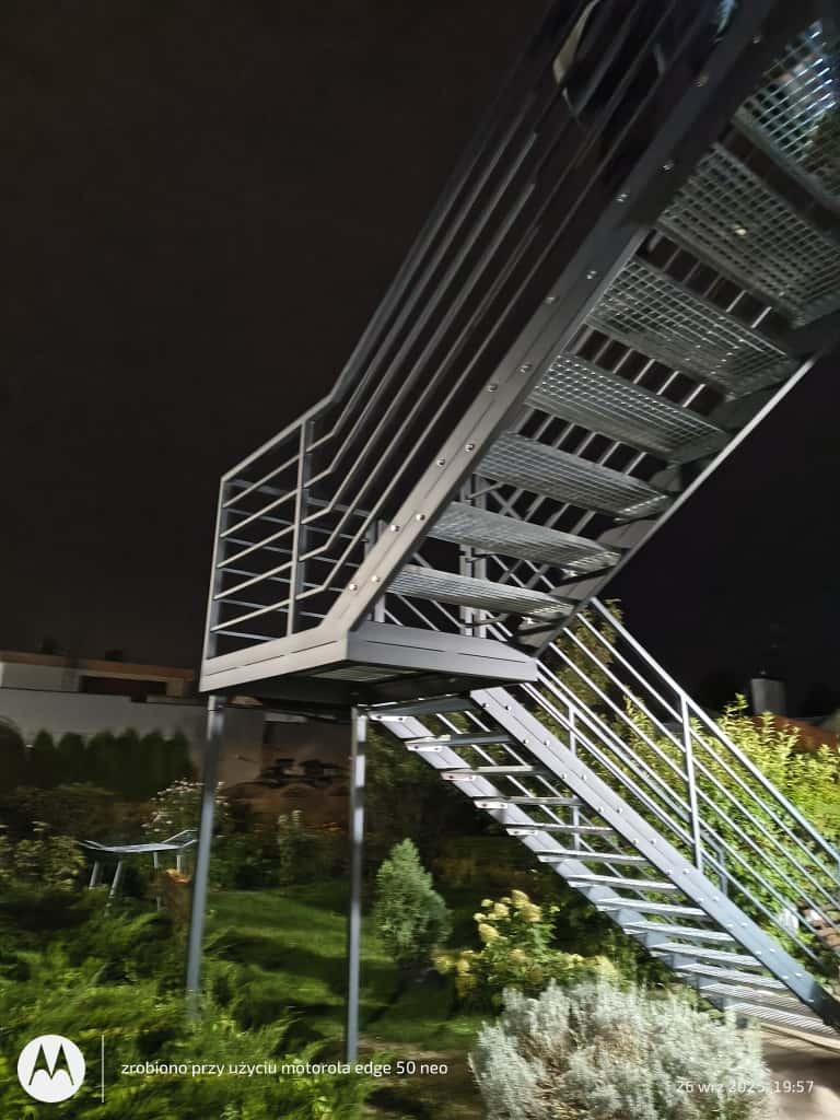 Zewnętrzne schody metalowe z ażurowymi stopniami, prowadzące do góry, z widocznym ogrodem w tle, sfotografowane nocą z dołu pod kątem.