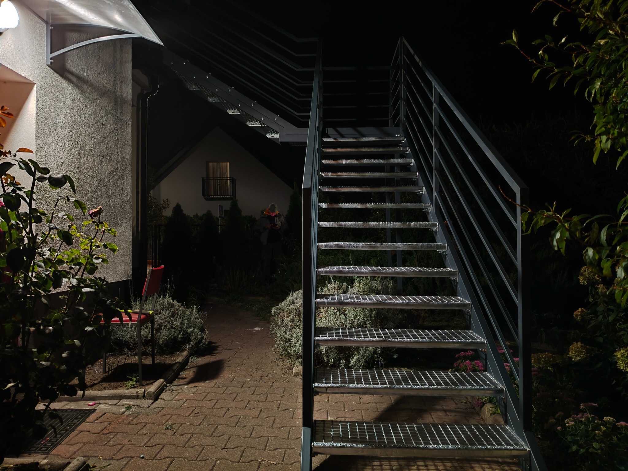 Zewnętrzne schody metalowe z ażurowymi stopniami prowadzące do domu, oświetlone nocą. Widoczne rośliny i ścieżka z kostki brukowej.