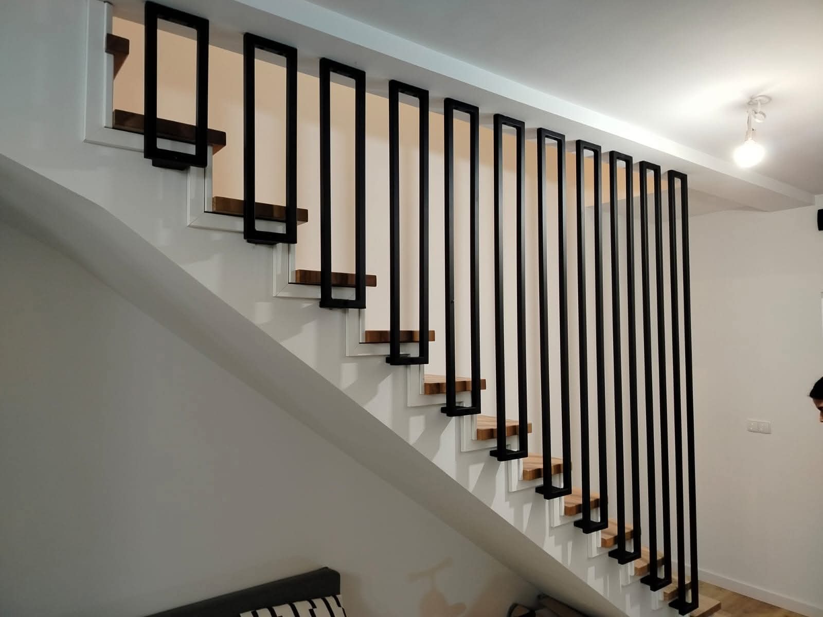 Nowoczesne schody z drewnianymi stopniami i czarną, minimalistyczną balustradą z pionowych, prostokątnych elementów. Białe ściany i sufit, widoczna lampa.