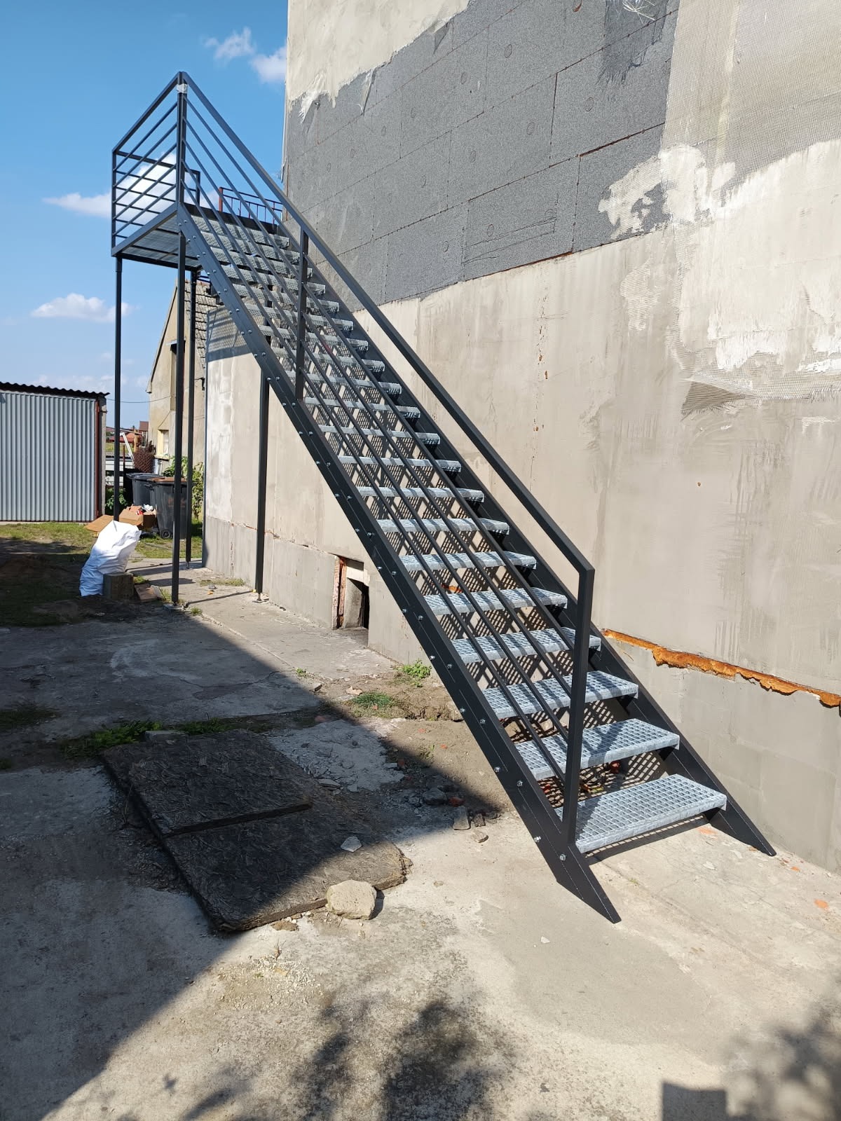 Zewnętrzne schody metalowe z platformą, prowadzące do budynku w trakcie remontu. Konstrukcja stalowa, ocynkowana, z ażurowymi stopniami. Widok z dołu, w tle budynek z ociepleniem.