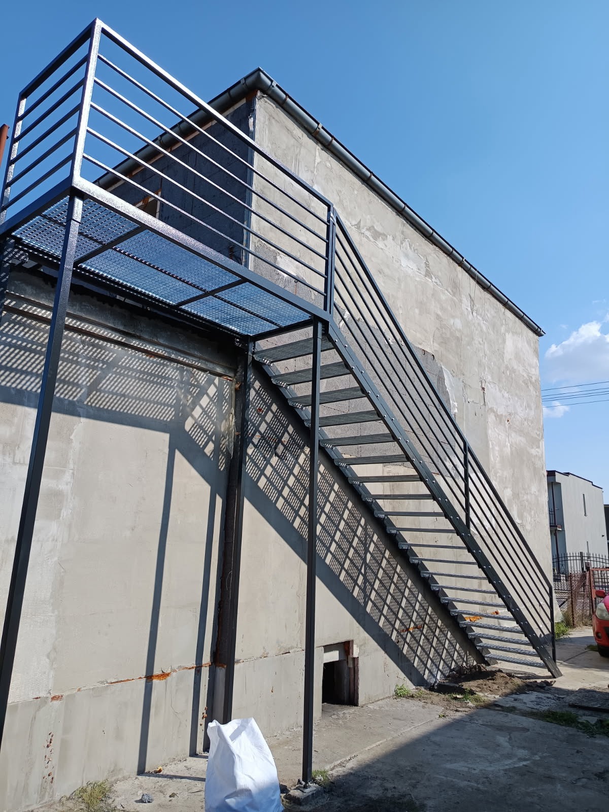 Zewnętrzne, metalowe schody z ażurową platformą i prostą balustradą, prowadzące do budynku o surowej, betonowej elewacji w słoneczny dzień. Cień schodów tworzy geometryczny wzór.