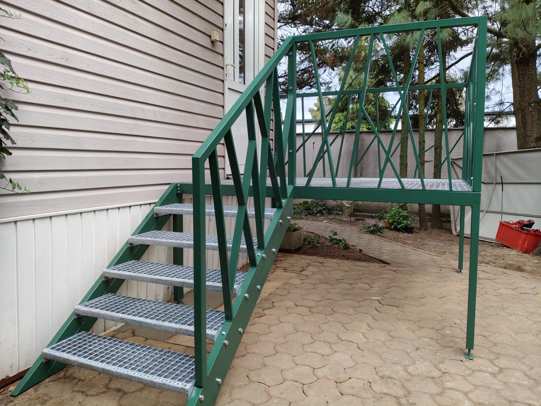 Zielone schody metalowe z ażurową balustradą prowadzą do drzwi domu z sidingiem. Widoczne mocowania śrubowe i ażurowa konstrukcja balustrady. Tło stanowią drzewa i kostka brukowa.