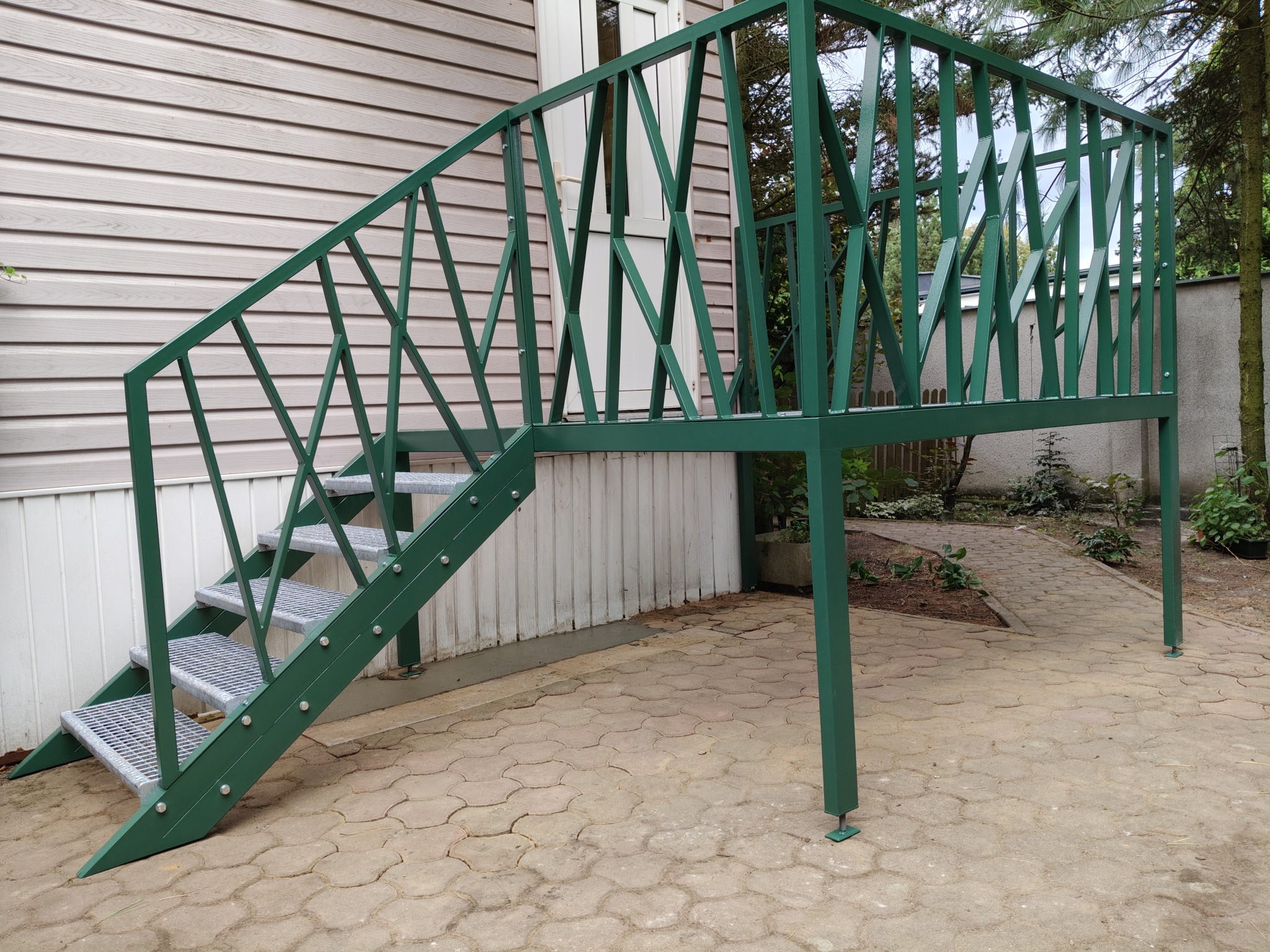 Zielone schody metalowe z ażurową balustradą, prowadzące do drzwi na podwyższeniu. Widoczne detale konstrukcyjne i otoczenie budynku.