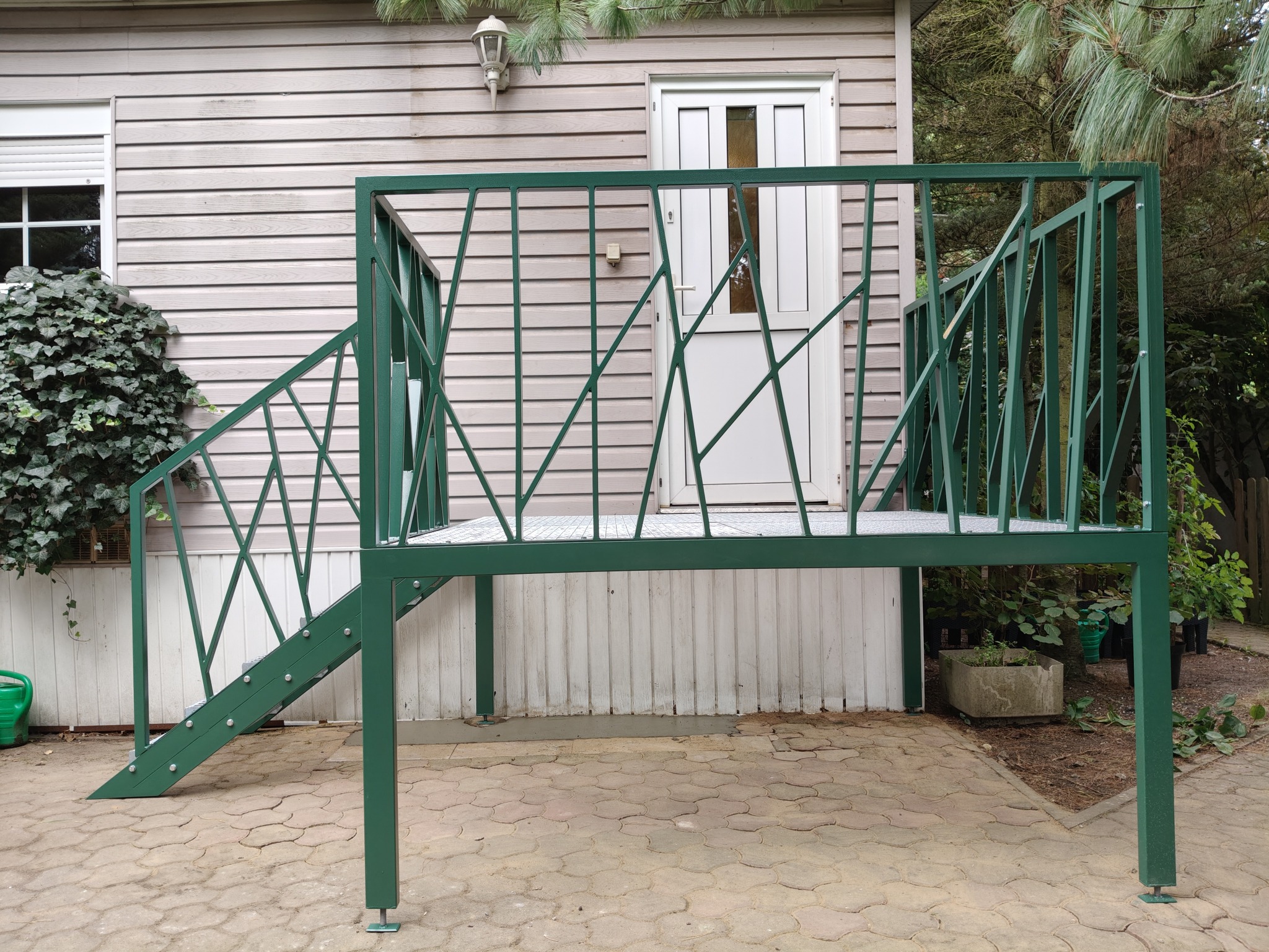 Zielona, metalowa konstrukcja schodów zewnętrznych z balustradą, prowadząca do drzwi domu z drewnianą elewacją. Nietypowy wzór balustrady z ukośnymi elementami.