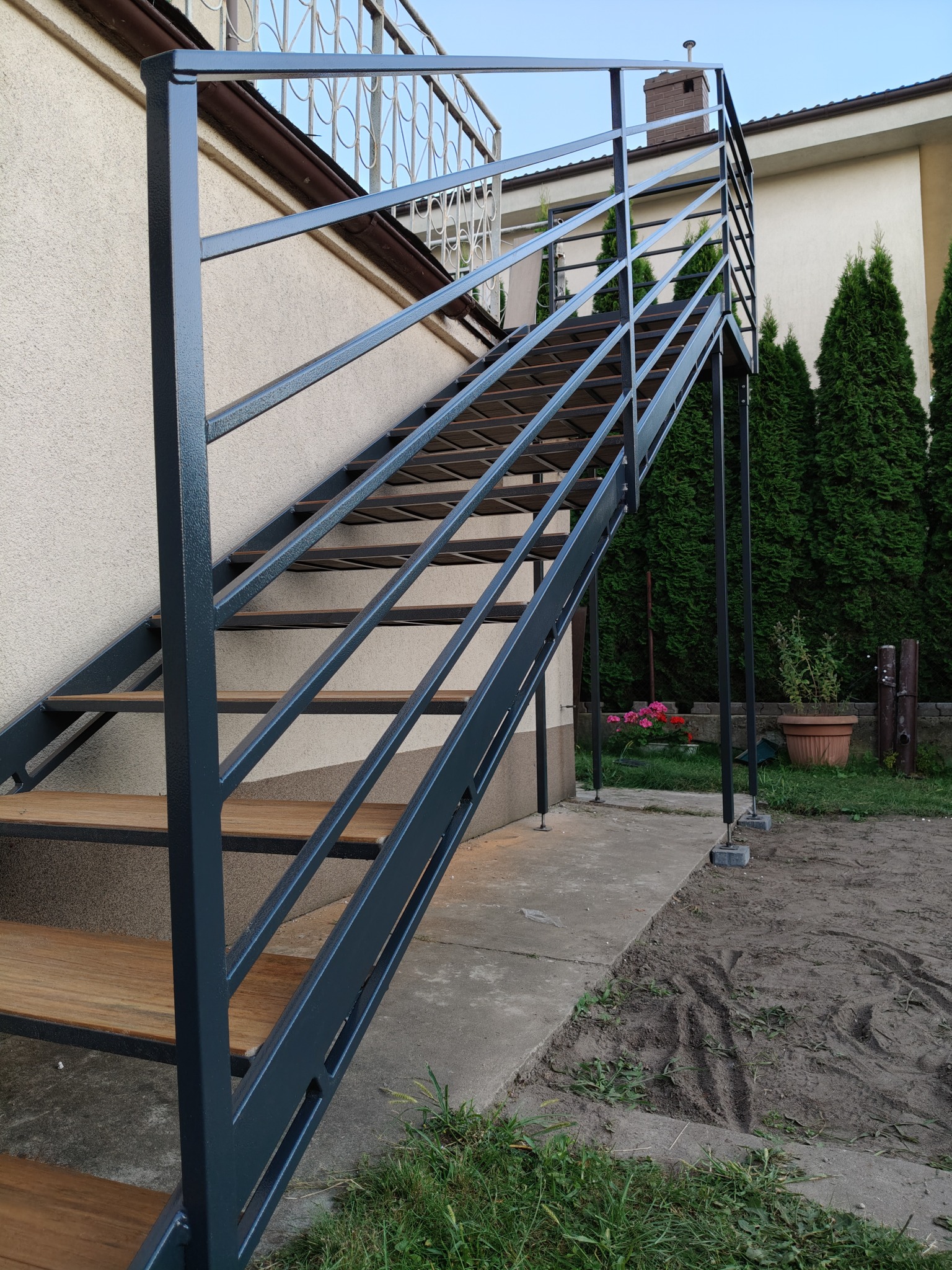 Zewnętrzne schody metalowe z drewnianymi stopniami, prowadzące na balkon. Konstrukcja w kolorze grafitowym, tło stanowią tuje i elewacja budynku.