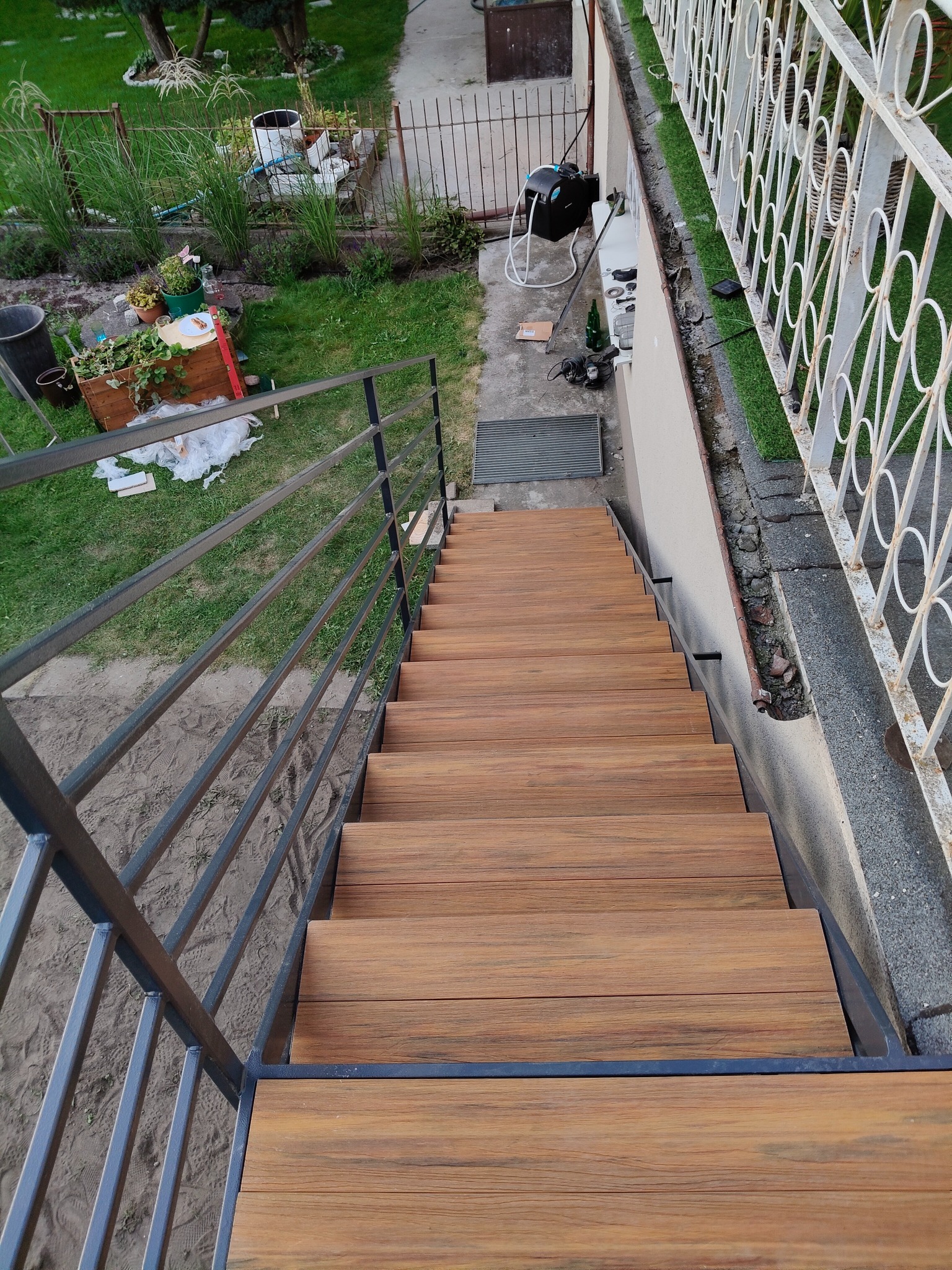 Drewnopodobne schody zewnętrzne z metalową balustradą, prowadzące w dół, z widokiem na ogród i elementy wyposażenia. Ujęcie z góry, podkreślające detale konstrukcji.