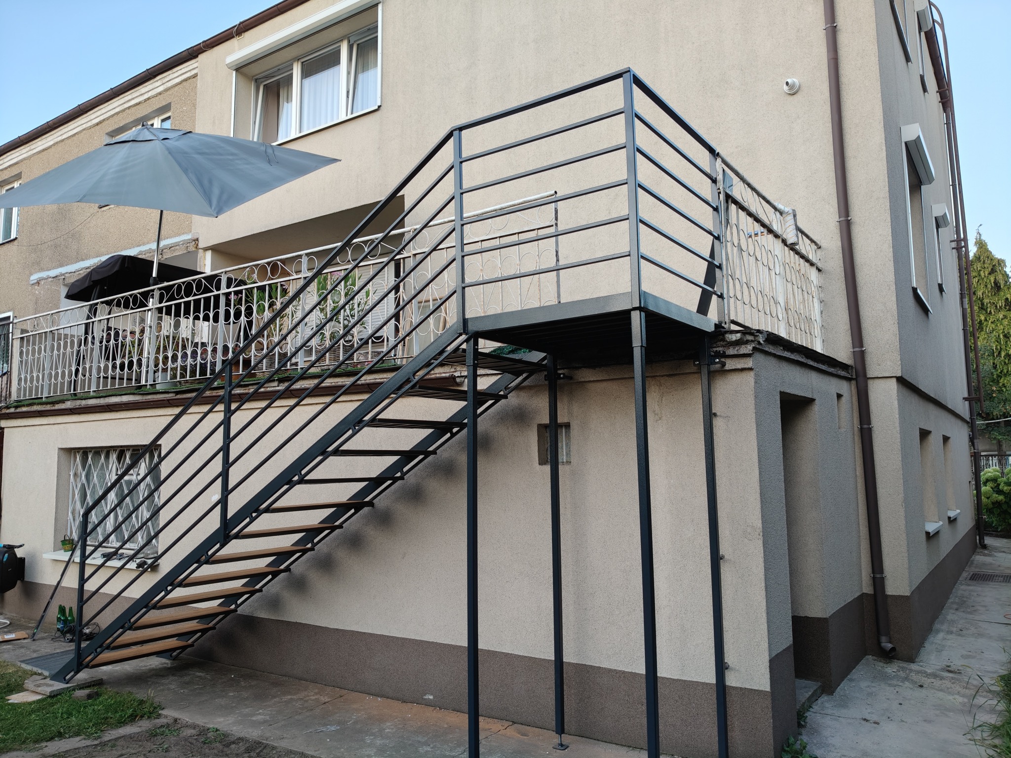 Zewnętrzne schody metalowe z drewnianymi stopniami, prowadzące na balkon w Mieścisko. Konstrukcja pomalowana na czarno, widok z dołu, jasne tło budynku.