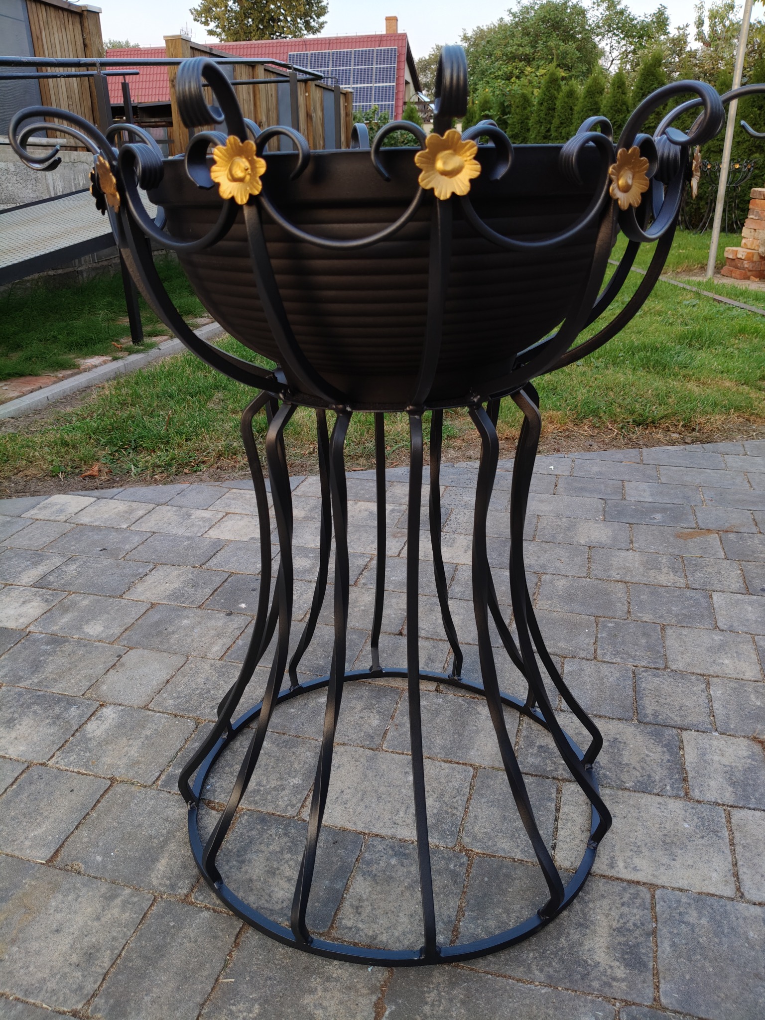 Kuta, czarna donica metalowa z dekoracyjnymi, złotymi kwiatami, stojąca na kostce brukowej. Widoczne elementy ogrodu i domu w tle.