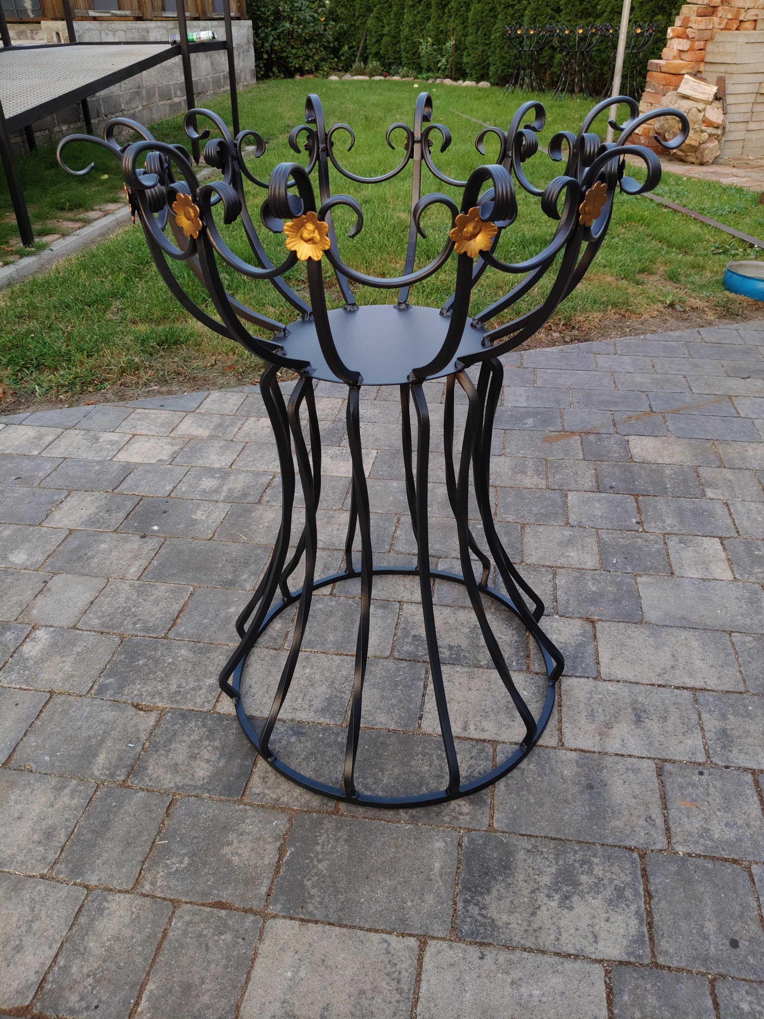 Kuta, czarna konstrukcja metalowa z dekoracyjnymi, złotymi kwiatami, stojąca na brukowanym podłożu w ogrodzie. Unikalny design i precyzja wykonania.