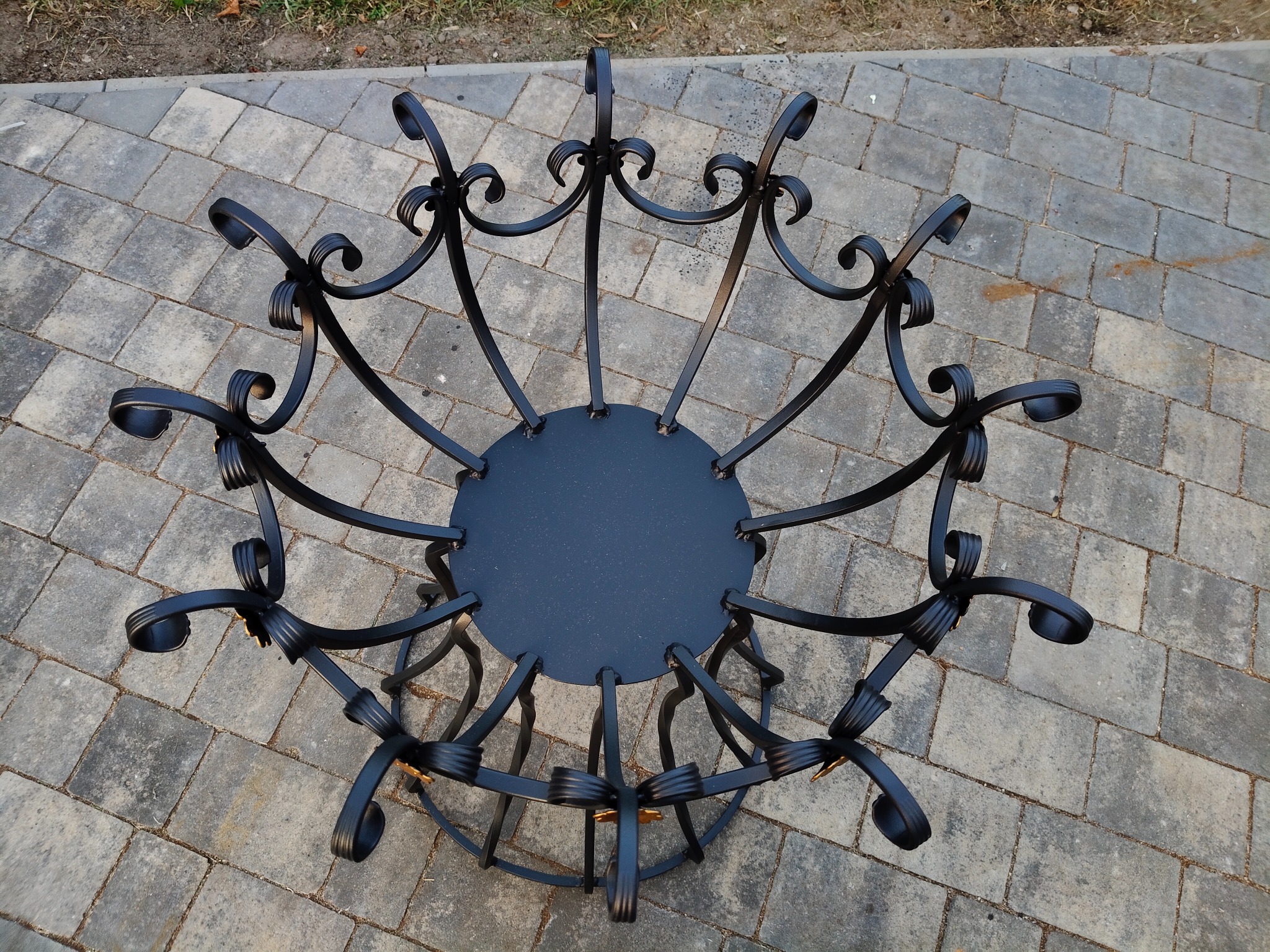 Kuta, czarna konstrukcja metalowa z dekoracyjnymi elementami, przypominająca kosz lub kwietnik, ustawiona na szarej kostce brukowej. Widok z góry.