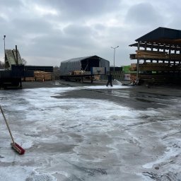 Michał Walczak M&W - Pracownik w ciemnym ubraniu spryskuje wodą pod ciśnieniem duży plac wyłożony kostką brukową, pokrytą częściowo pianą czyszczącą; w tle składowane drewno, hala namiotowa i pochmurne niebo.