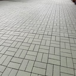 Michał Walczak M&W - Szara kostka brukowa ułożona we wzór jodełki na podjeździe. Widok z perspektywy, z garażem i zielenią w tle. Układanka z detalami.