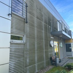 Michał Walczak M&W - Zabrudzona, pofałdowana elewacja budynku z metalu, częściowo pokryta zielonym nalotem. Widoczne okna, balkon z barierką i tablica informacyjna.