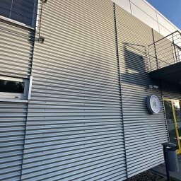 Michał Walczak M&W - Fasada budynku z blachy falistej w odcieniach szarości, z widocznym oknem, balkonem, tablicą informacyjną i koszem na śmieci. U dołu widoczne kamienie.