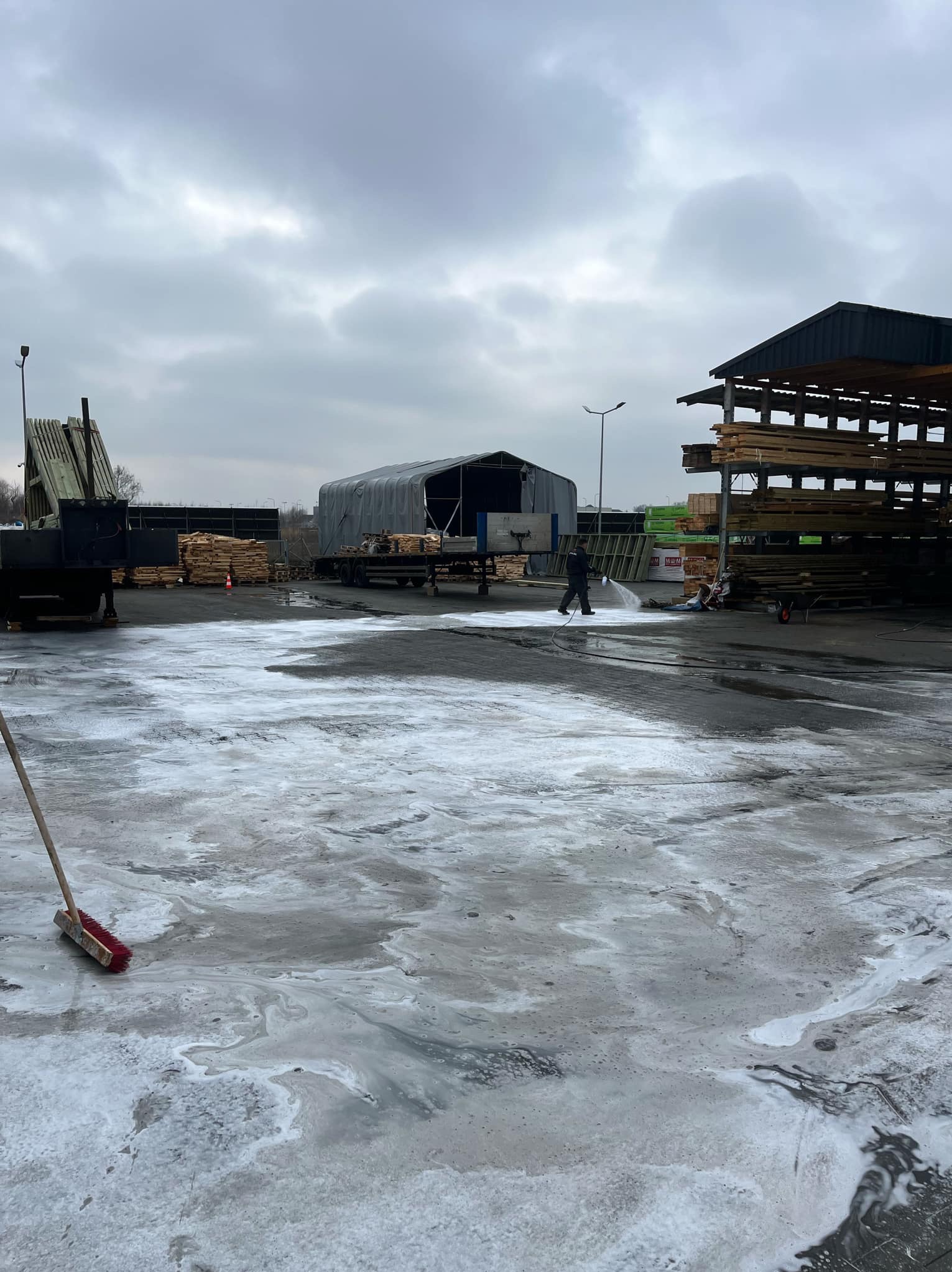 Pracownik w ciemnym ubraniu spryskuje wodą pod ciśnieniem duży plac wyłożony kostką brukową, pokrytą częściowo pianą czyszczącą; w tle składowane drewno, hala namiotowa i pochmurne niebo.