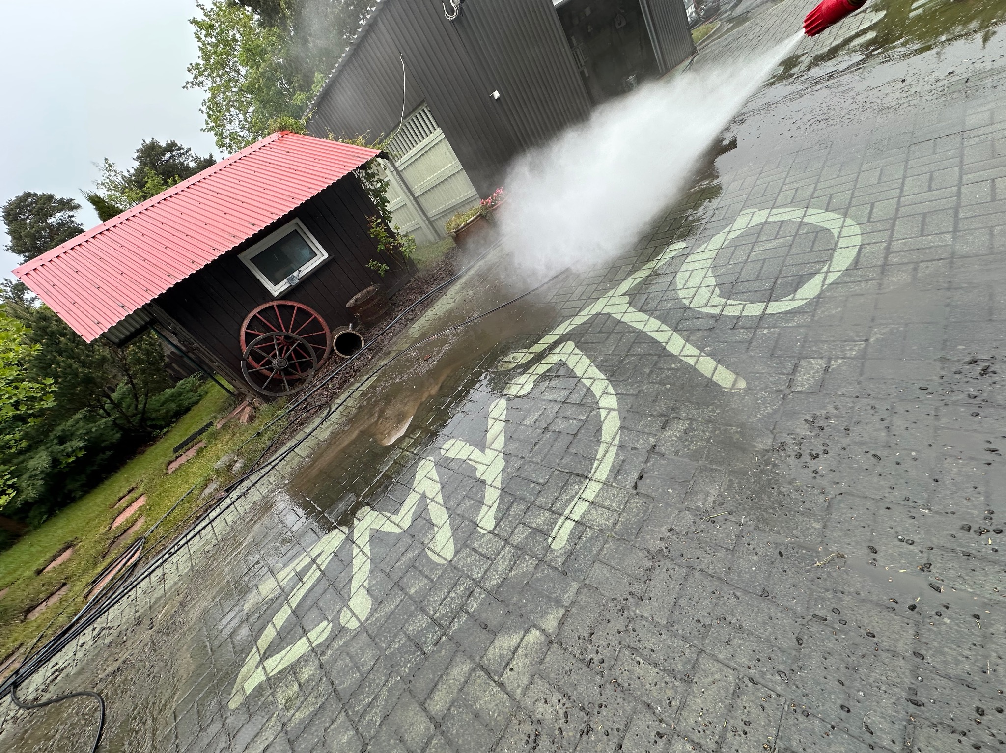 Kostka brukowa czyszczona myjką ciśnieniową, na której widać wzór. W tle domek narzędziowy z czerwonym dachem i budynek gospodarczy. Ujęcie z góry, lekko pod kątem.