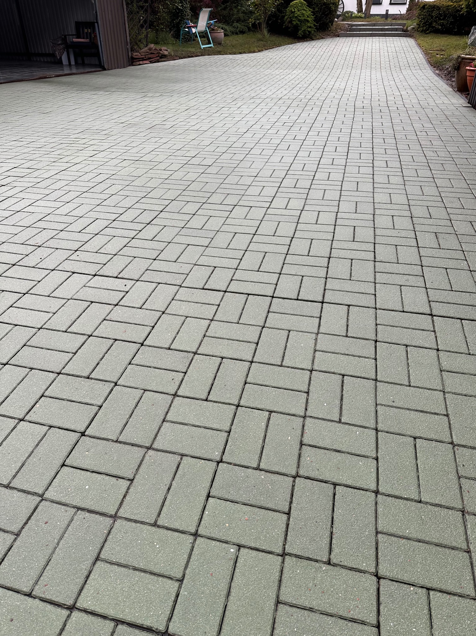 Szara kostka brukowa ułożona we wzór jodełki na podjeździe. Widok z perspektywy, z garażem i zielenią w tle. Układanka z detalami.