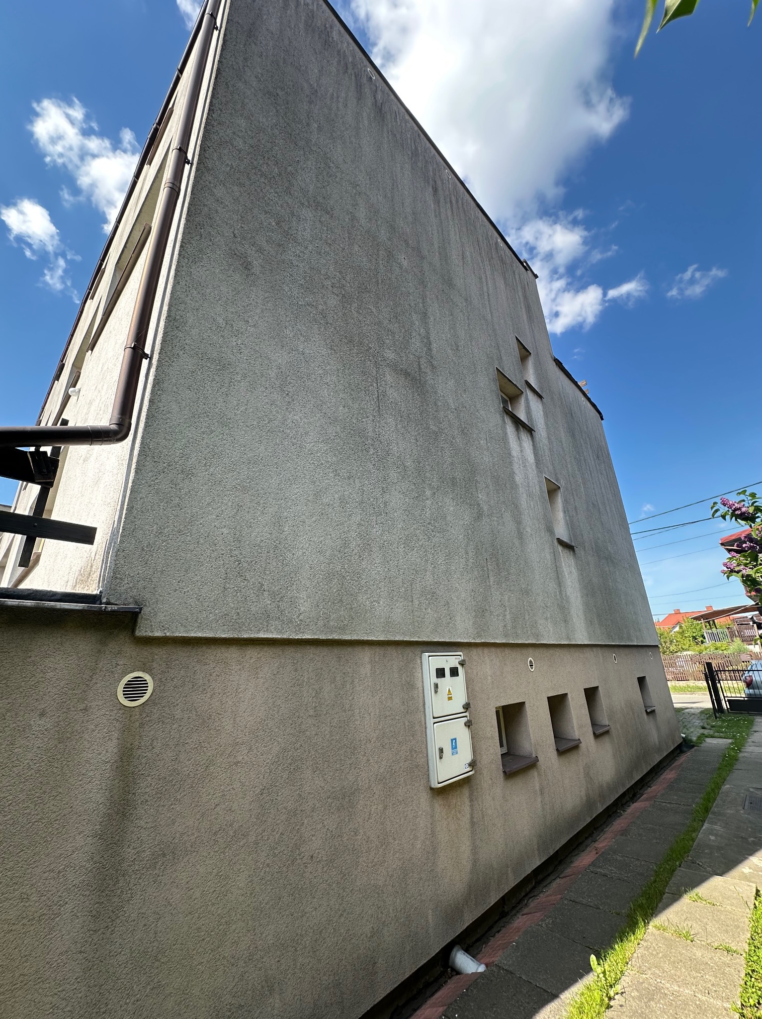 Szara, kilkupiętrowa elewacja budynku z rynnami w Tuszynie, widziana z dołu pod kątem. Widoczne małe okna i skrzynka elektryczna na ścianie.