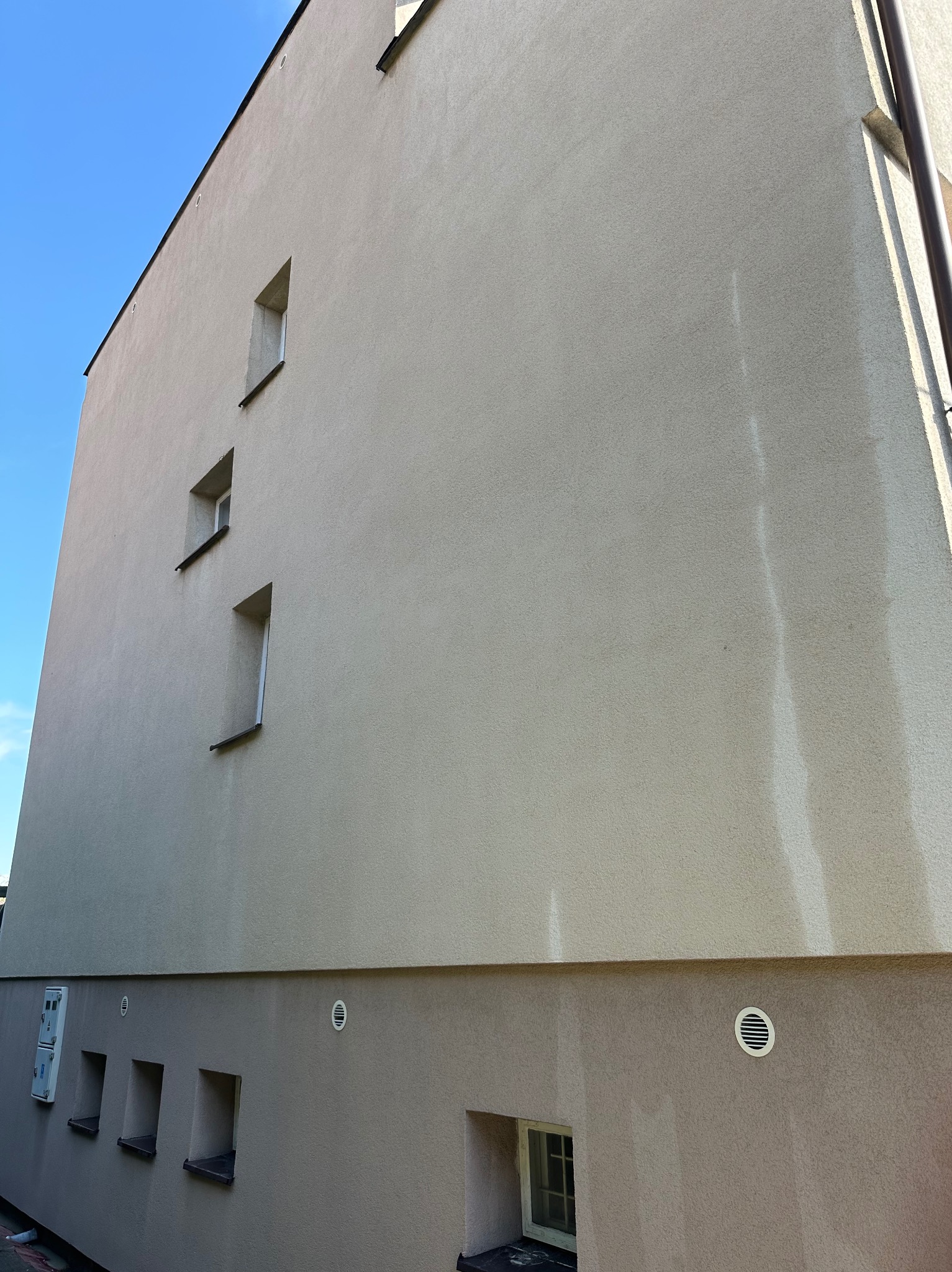 Ściana budynku z jasną elewacją, widoczne okna o nietypowym kształcie i ślady po umyciu. Część elewacji jest wyraźnie jaśniejsza, co sugeruje niedawne czyszczenie.