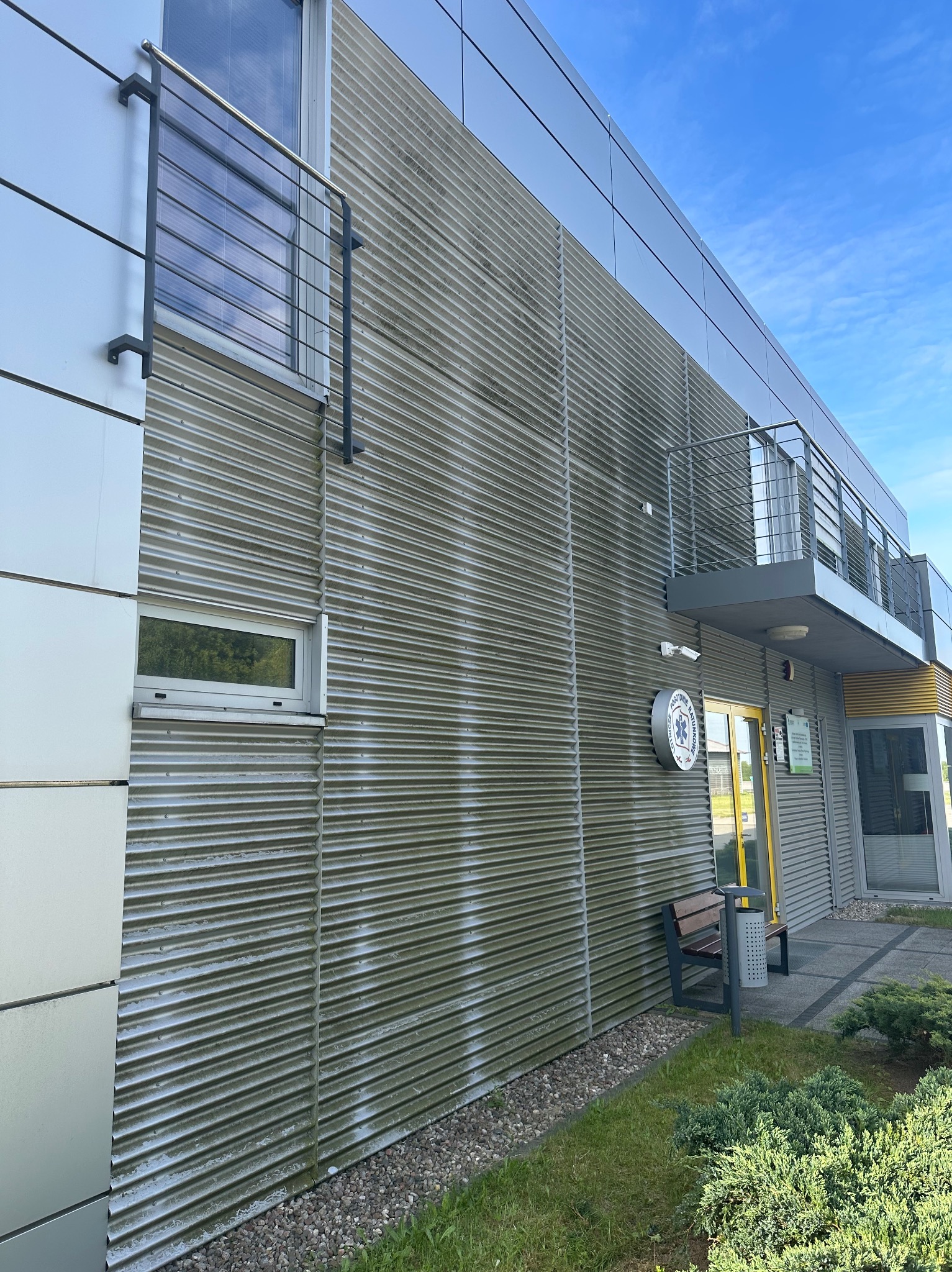 Zabrudzona, pofałdowana elewacja budynku z metalu, częściowo pokryta zielonym nalotem. Widoczne okna, balkon z barierką i tablica informacyjna.