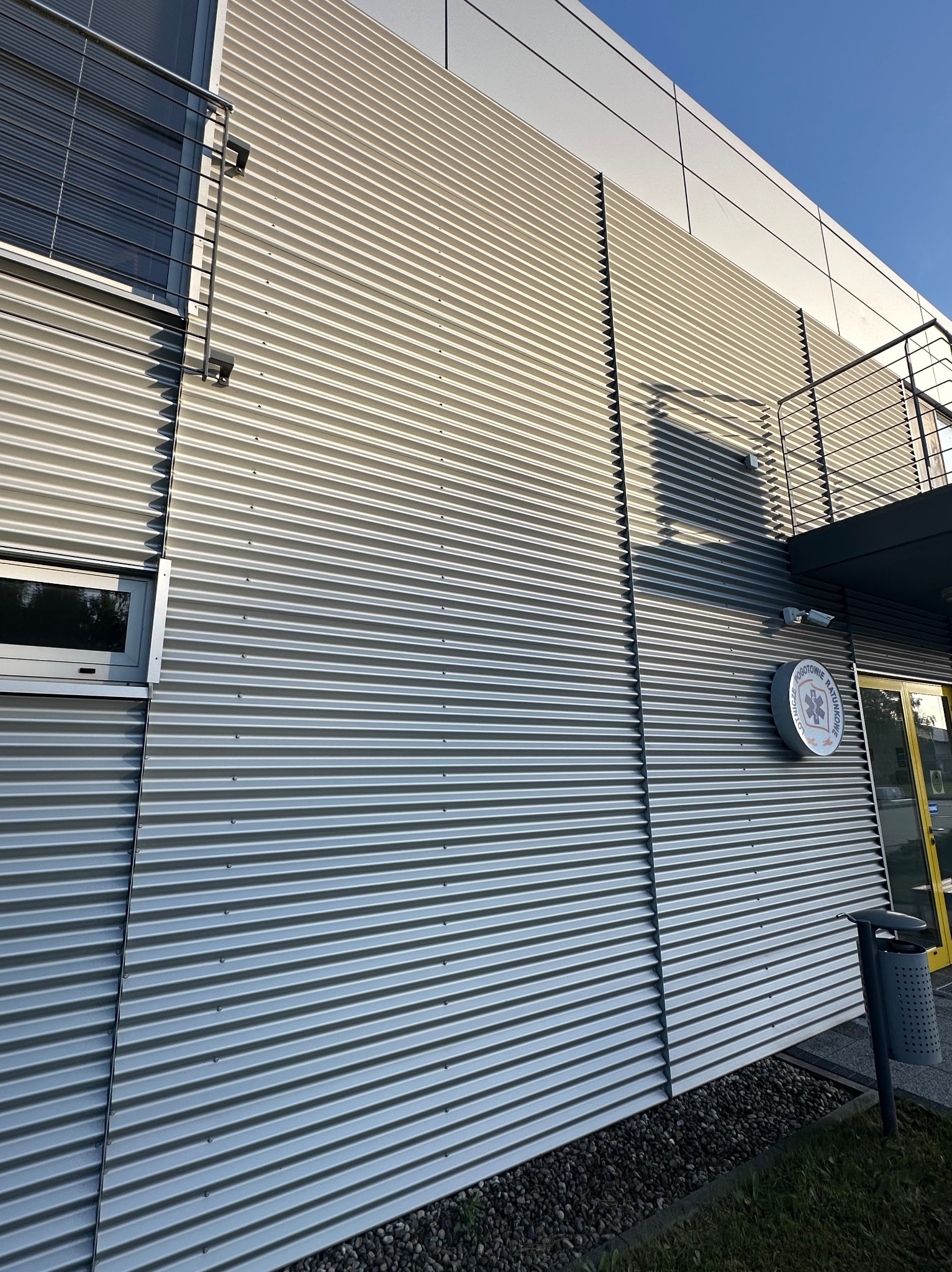 Fasada budynku z blachy falistej w odcieniach szarości, z widocznym oknem, balkonem, tablicą informacyjną i koszem na śmieci. U dołu widoczne kamienie.