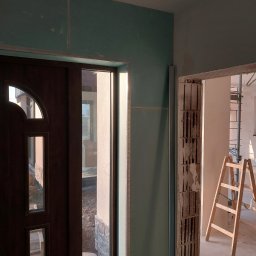 REMLUX Krzysztof Kozłowski - Montaż płyt karton gips na ścianie przy drzwiach wejściowych, widoczna surowa cegła w przejściu do kolejnego pomieszczenia, drabina oparta o ścianę, widok z dołu.