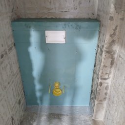 Niebieska płyta karton-gips zamontowana na ścianie w surowej łazience, widoczne żółte elementy instalacji. Ściany i podłoga w stanie deweloperskim.