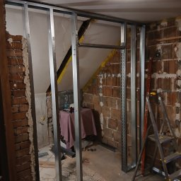 Konstrukcja ścianki działowej z profili stalowych na tle surowej cegły i fragmentu poddasza. Widoczne narzędzia i drabina. Etap prac wykończeniowych.