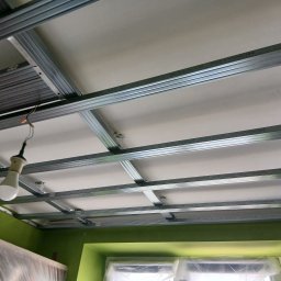 Sufit z płyt kartonowo-gipsowych w trakcie montażu, widoczne metalowe profile i częściowo zamontowane płyty. W tle zielona ściana i okno z folią ochronną.