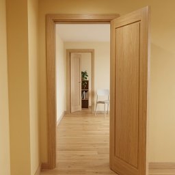 REMLUX Krzysztof Kozłowski - Jasne wnętrze z drewnianą podłogą i otwartymi drzwiami, przez które widać dalszą część mieszkania z regałem i krzesłem. Ciepła kolorystyka ścian i naturalne światło.