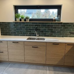 REMLUX Krzysztof Kozłowski - Nowoczesna kuchnia z drewnianymi szafkami, białym blatem i zielonymi płytkami na ścianie. Nad blatem okno z widokiem na zewnątrz. Minimalistyczny design.