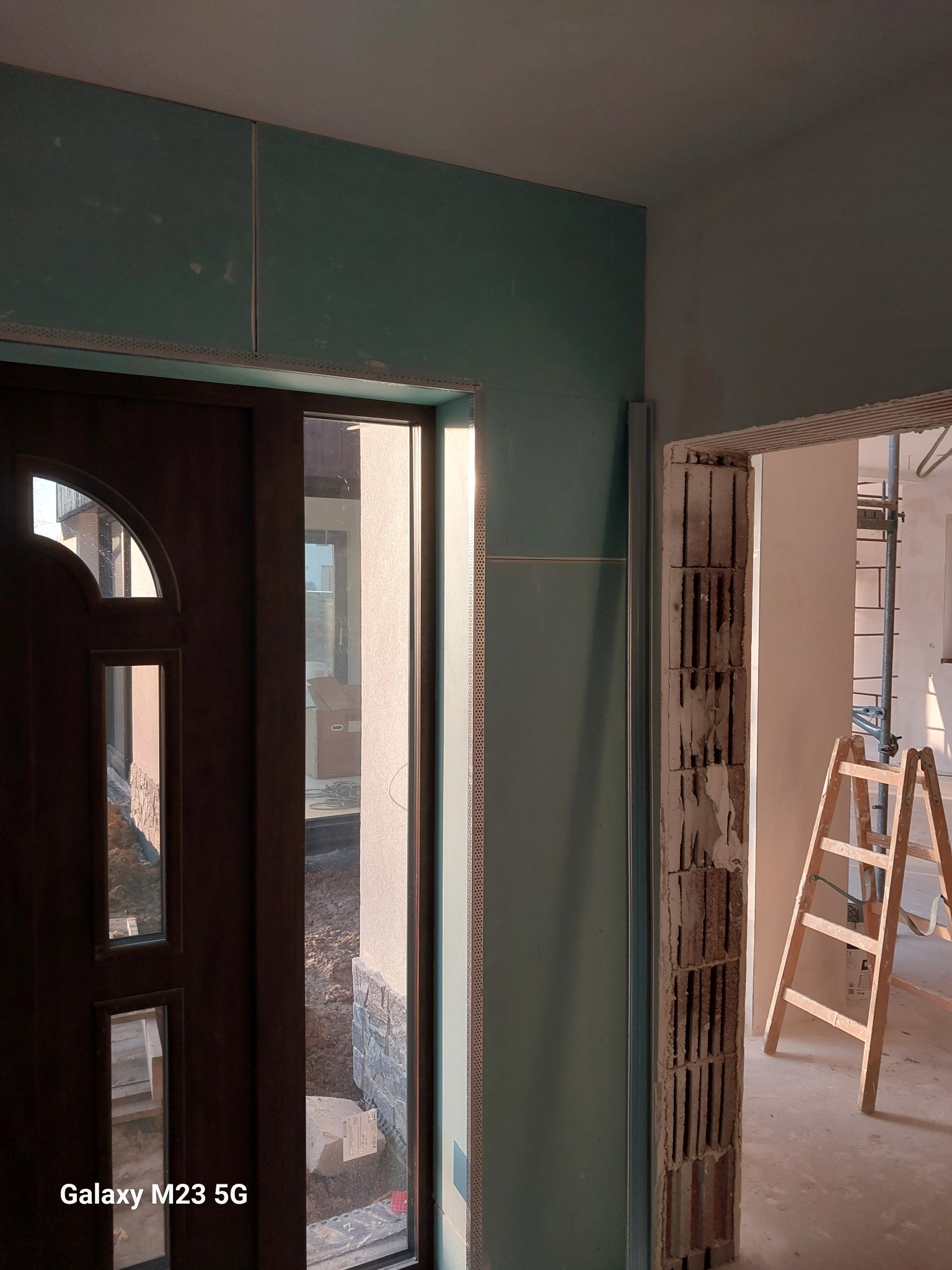 Montaż płyt karton gips na ścianie przy drzwiach wejściowych, widoczna surowa cegła w przejściu do kolejnego pomieszczenia, drabina oparta o ścianę, widok z dołu.