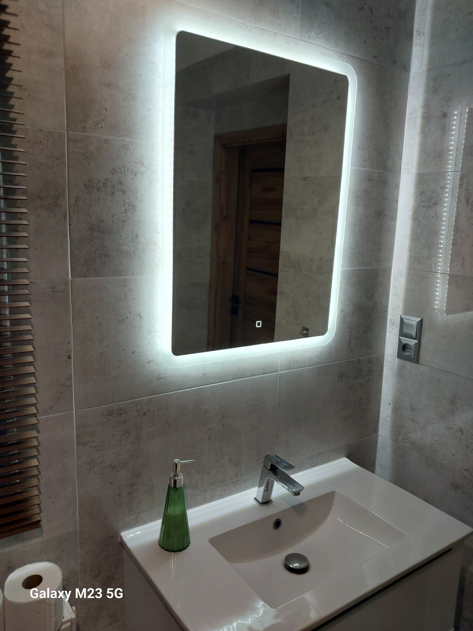 Nowoczesna łazienka z umywalką nablatową, podświetlanym lustrem LED i szarymi płytkami imitującymi kamień, widoczny dozownik mydła w kształcie stożka i drewniana żaluzja.